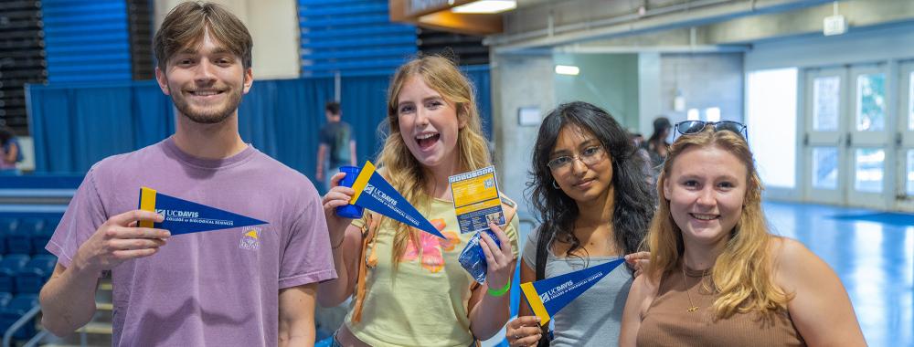 Four students smile and hold small UC Davis pennants during a campus event inside a large indoor space
