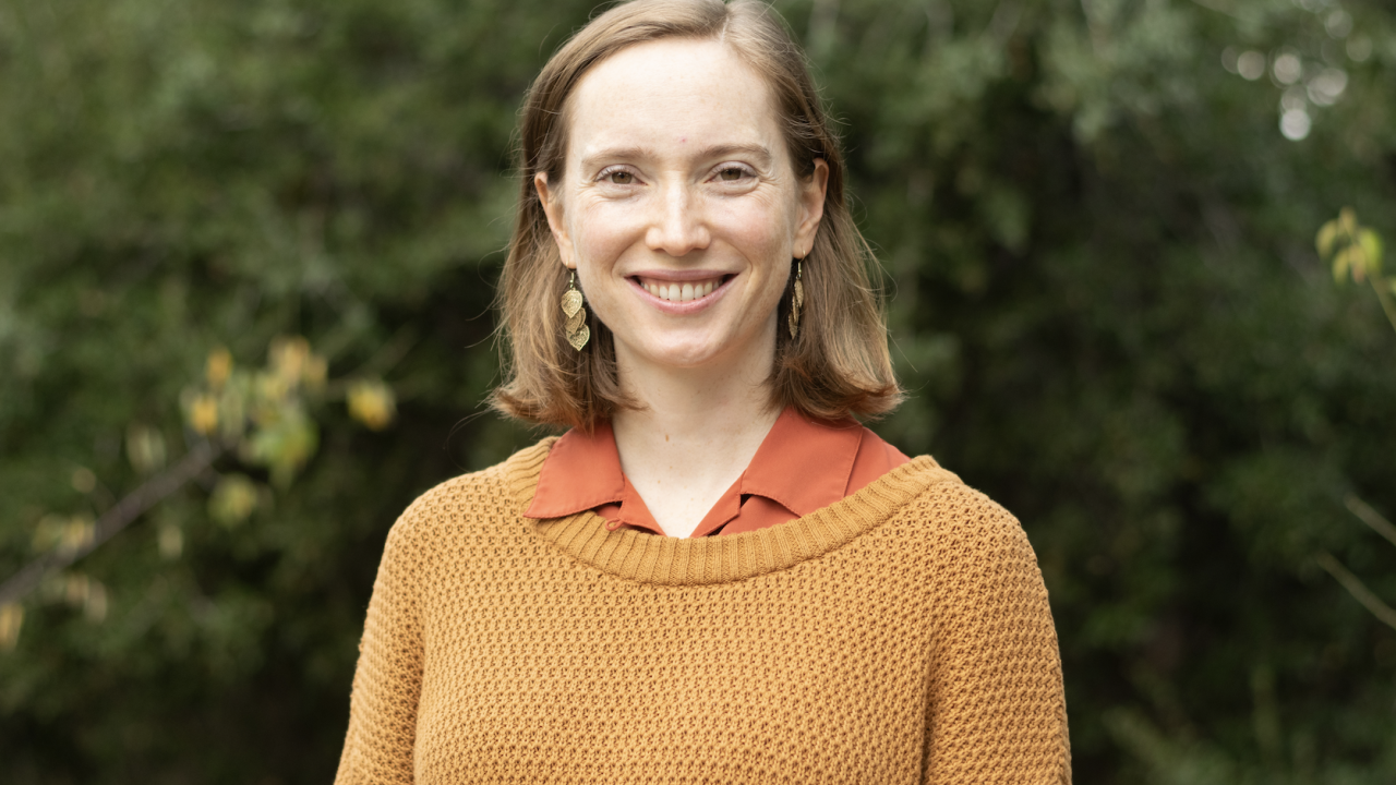 Person smiling outdoors in front of greenery, wearing a mustard knit sweater over a rust-colored collared shirt.