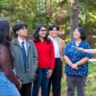 Instructor gestures while talking with smiling students gathered in the arboretum surrounded by trees and greenery.